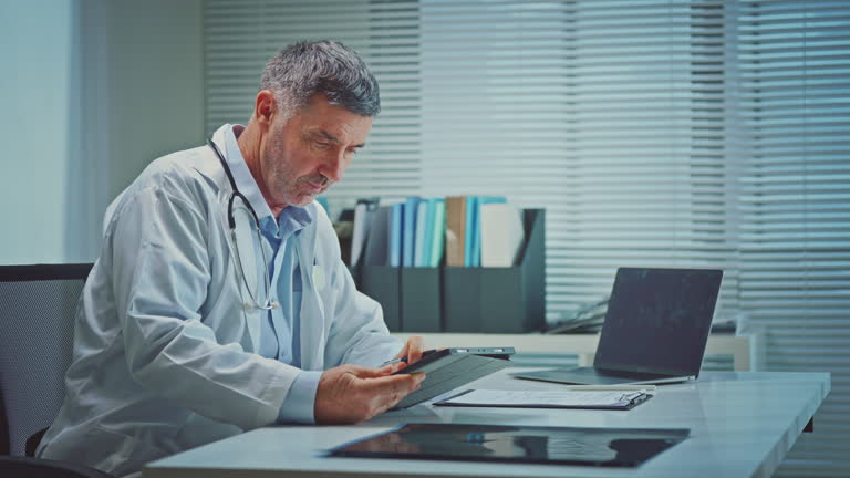 A male doctor in a white coat attentively reviews medical records on a tablet in a modern office.